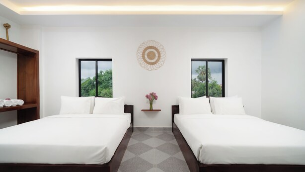 A hotel room with two beds, a small shelf with towels, a wall-mounted decorative item, and a view of greenery through the windows. - Visak Pool Villa  (Siem Reap)