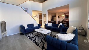 Un salón amplio con dos sofás azules, una mesa de centro blanca y un tapete de diseño blanco y negro. Una escalera conduce a un nivel superior.
