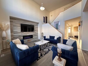 A modern living room with a flat-screen TV, a fireplace, blue sofas, and a staircase leading to another level.