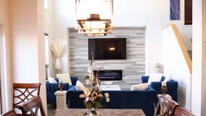 Una sala de estar moderna con un televisor de pantalla plana grande montado en una pared gris, un sofá azul, una mesa de comedor con sillas y un jarrón decorativo sobre la mesa.