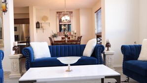 Una sala de estar con un sofá azul, una mesa de centro blanca y un comedor al fondo.
