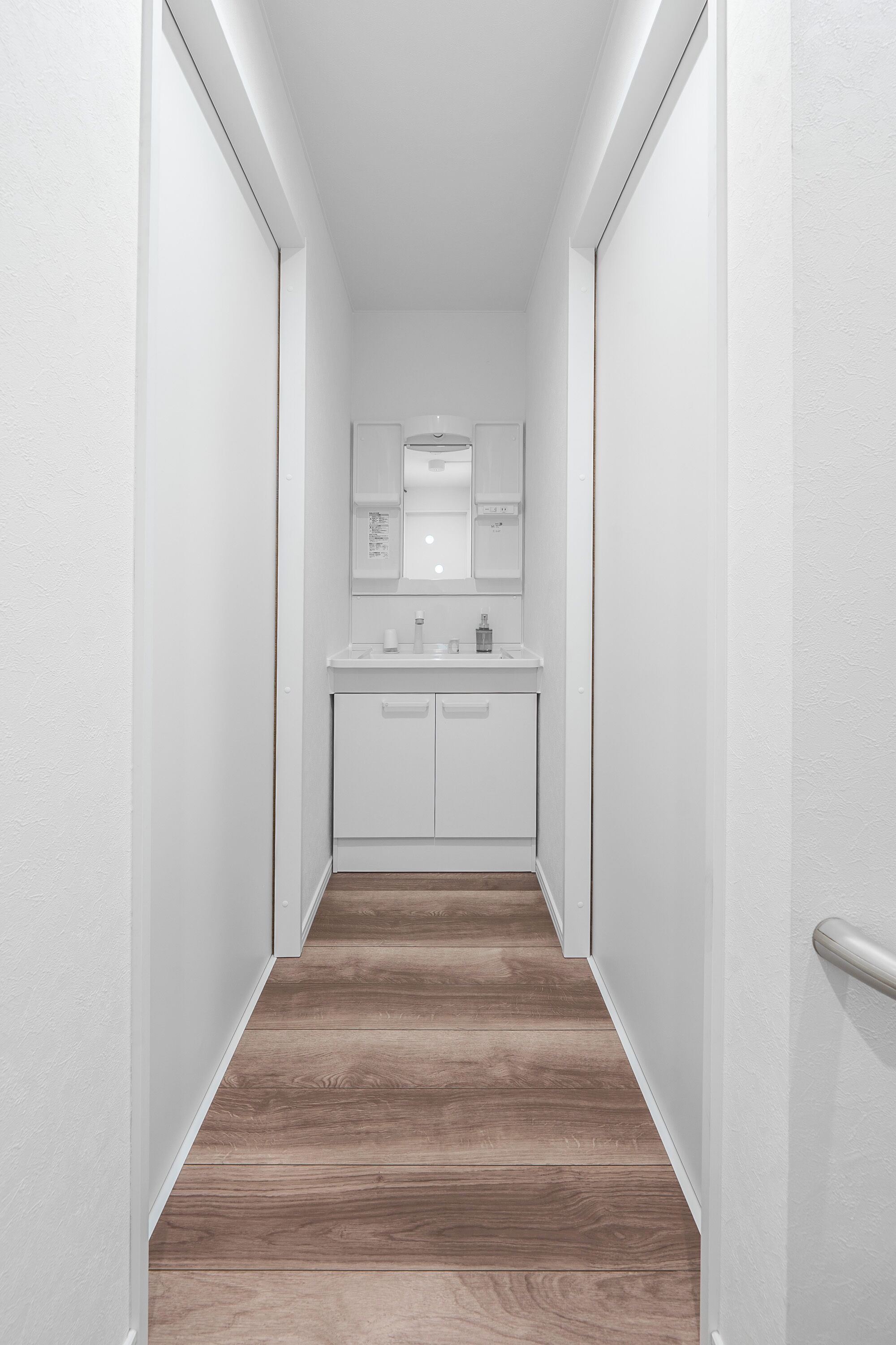Un couloir étroit, doté d’un plancher en bois, mène à une salle de bain avec un lavabo et un miroir blancs.