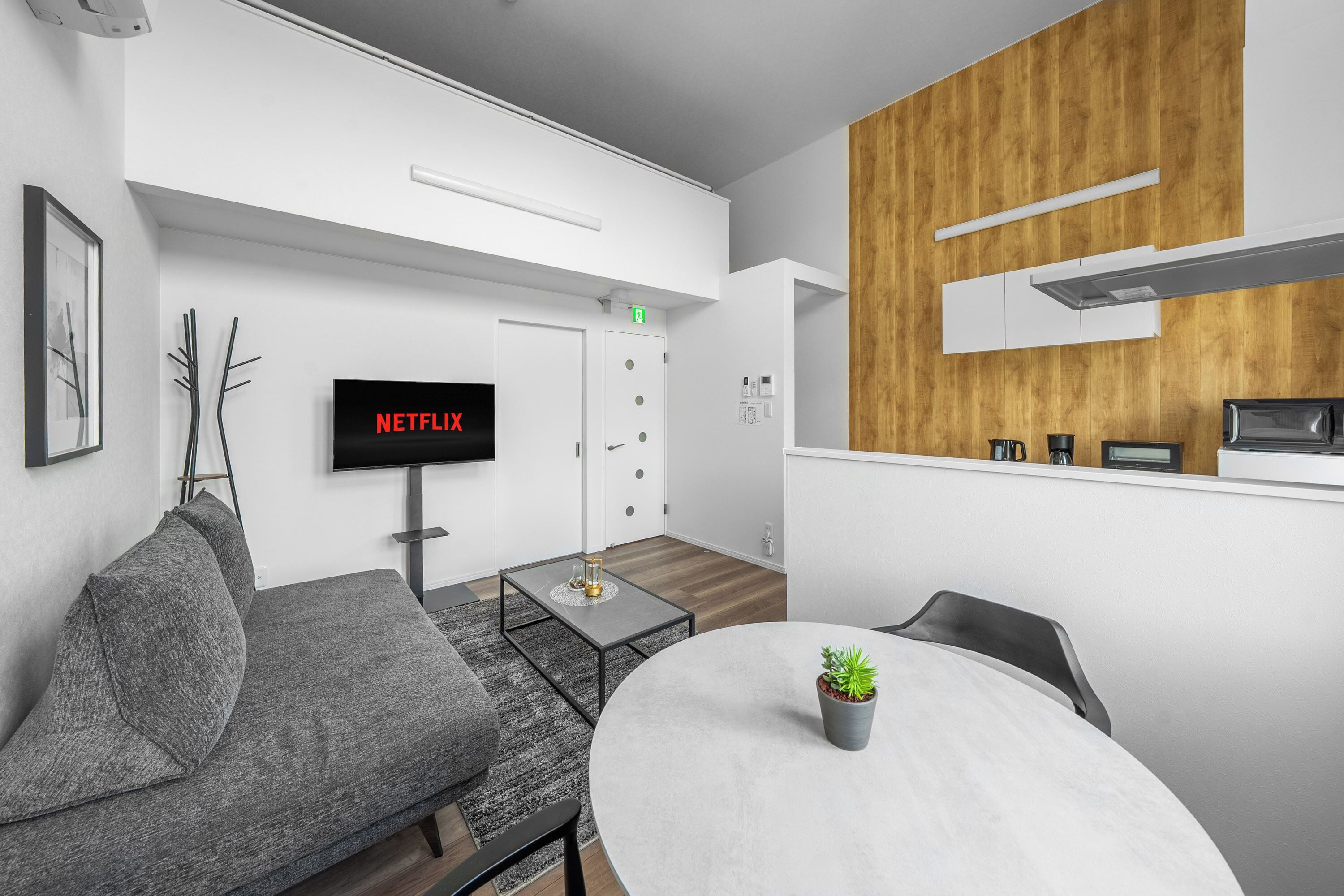 A modern living room with a grey sofa, a round table with a potted plant, a flat-screen TV displaying 'NETFLIX', and a wooden accent wall.