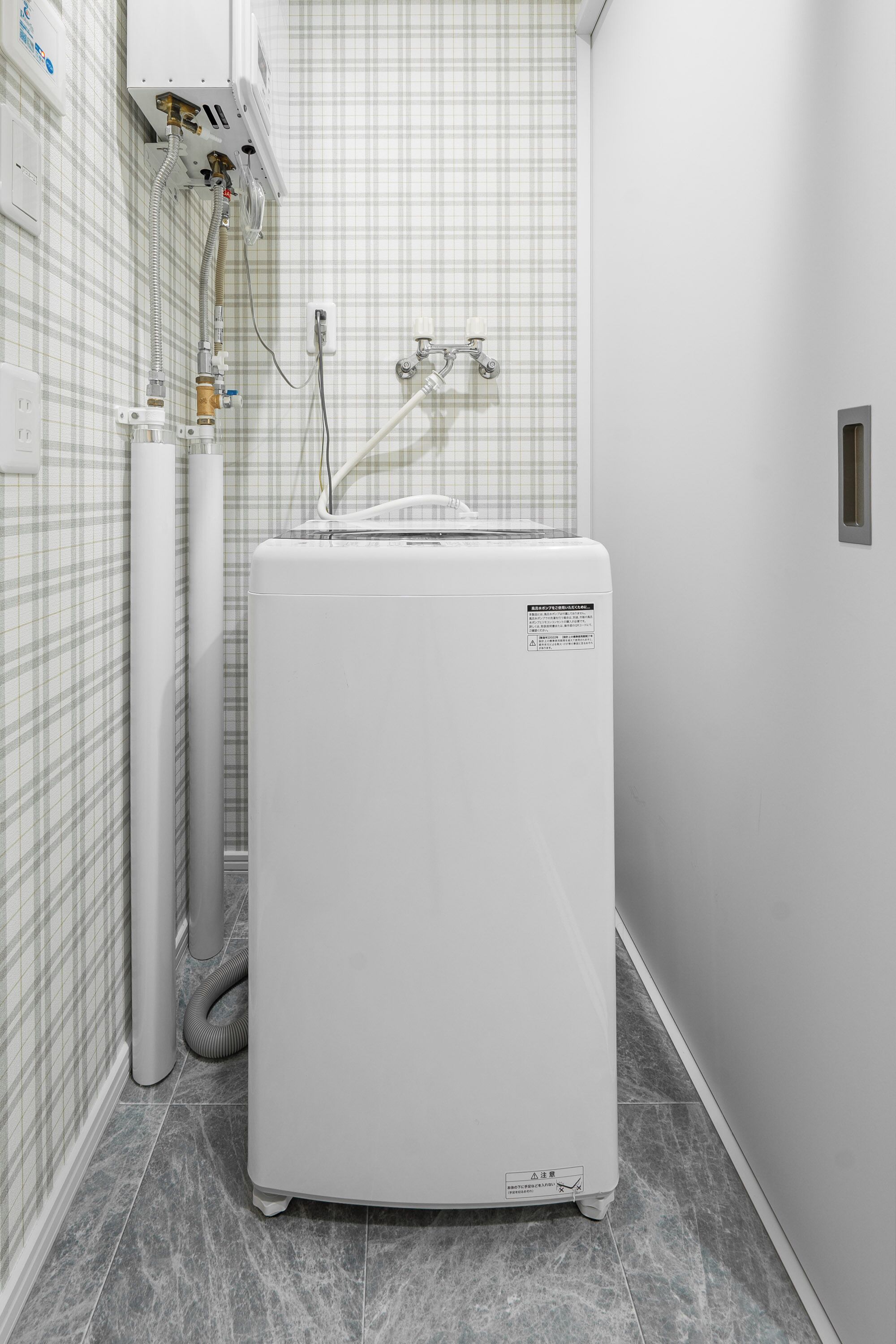 A compact, white, wall-mounted water heater with a control panel, connected to a flexible hose and a metal pipe, against a checkered wall.