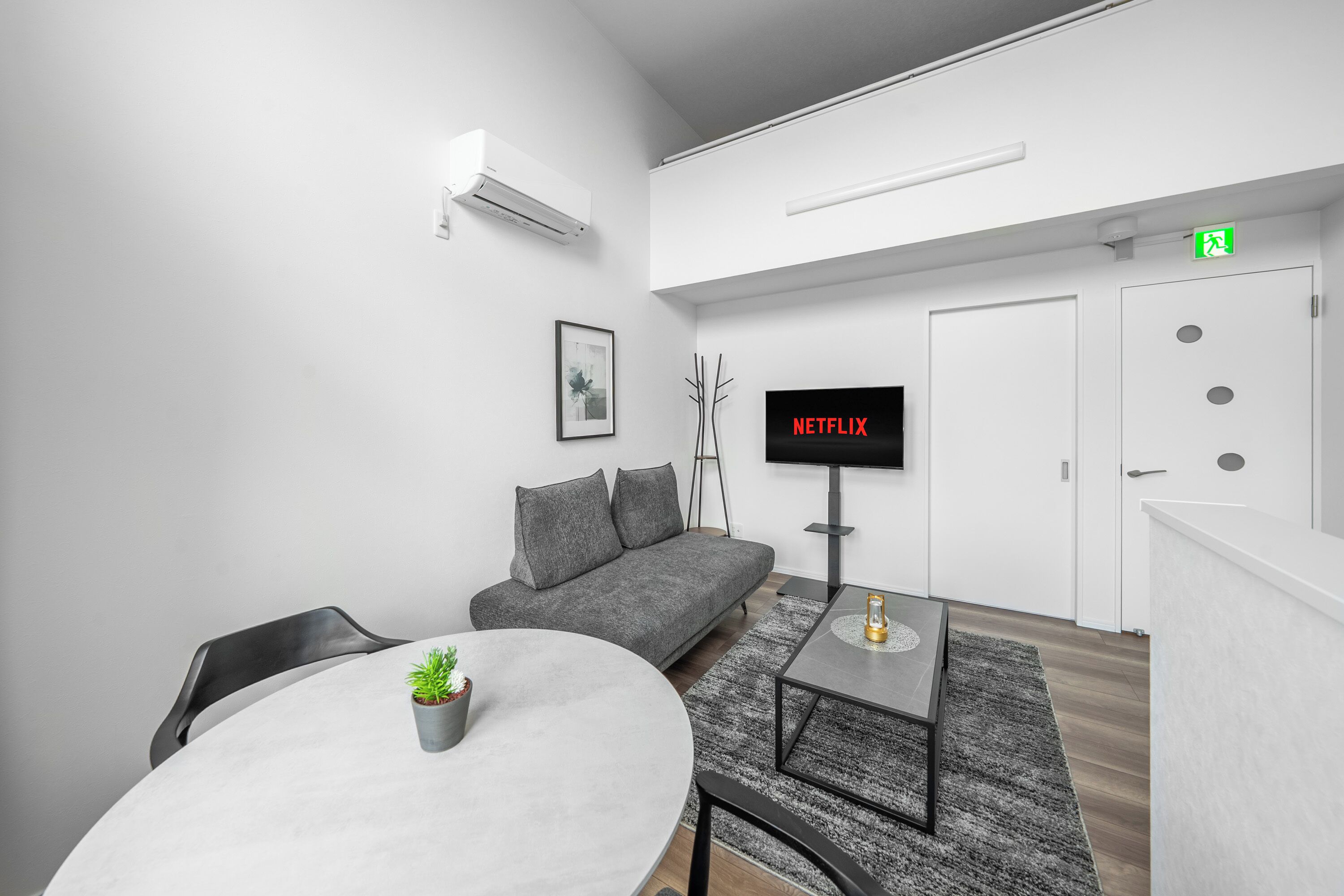 A modern living room with a grey sofa, a round table with a potted plant, a flat-screen TV displaying 'NETFLIX', and a staircase leading to an upper level.