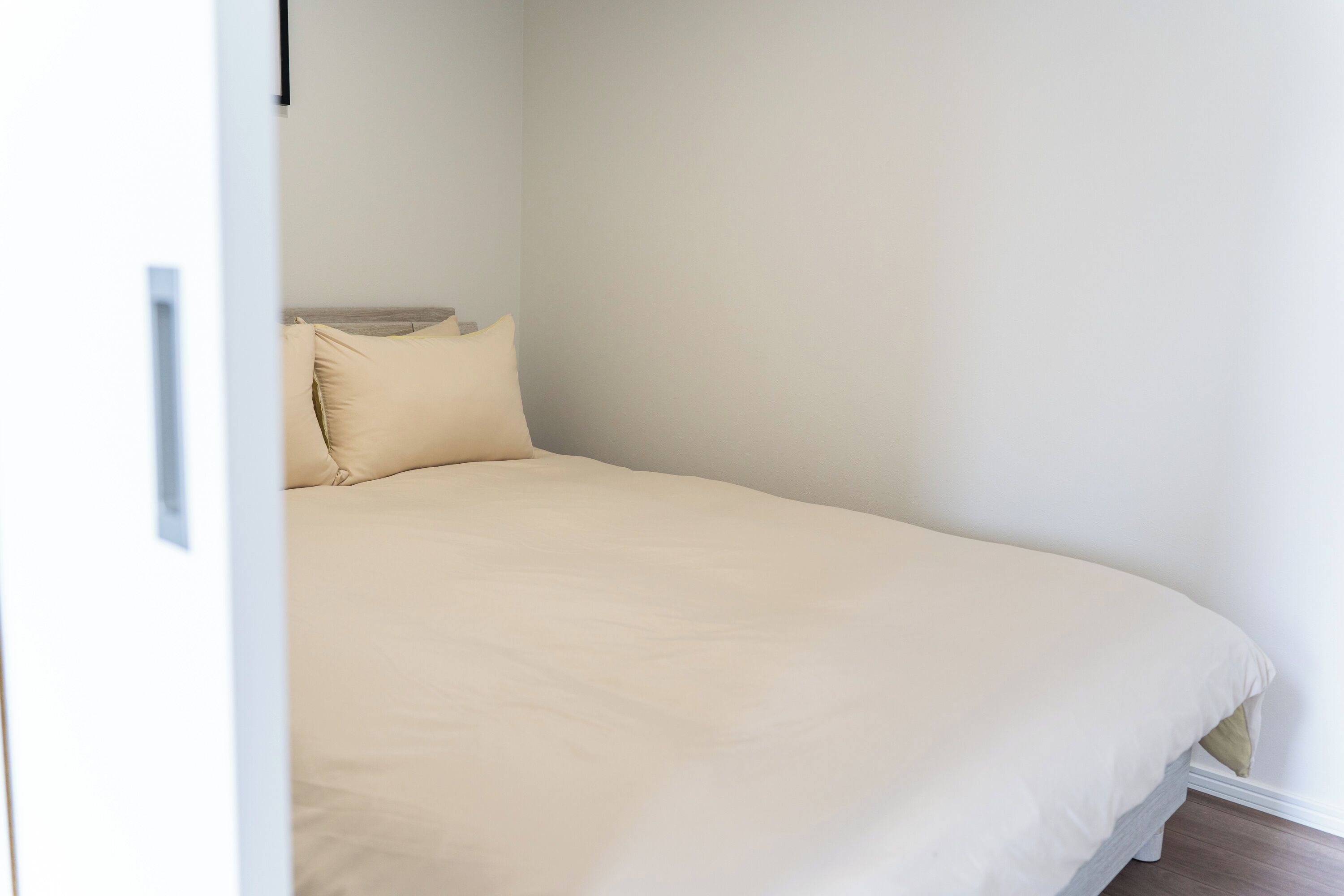 A single bed with a beige mattress and pillows, a white wall, and a wooden floor.