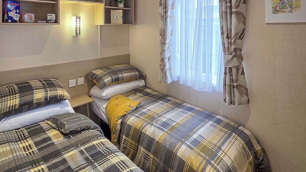A room with two beds, a shelf with decorative items, a window with sheer curtains, and a wall-mounted light.