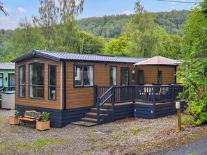 A wooden cabin with a balcony and a small deck, surrounded by gravel and greenery. - 2 bedroom accommodation in Birnam (Birnam)
