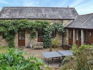 A stone building with a sloped roof, a wooden door, and a small garden area with a ping pong table. - 3 bedroom accommodation in Liskeard (Liskeard)