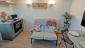 A modern kitchen with a built-in stove, a dining area with a round table and chairs, a sofa with floral cushions, and a wall-mounted light fixture. - Raygill Shepherds Hut (Dishforth, near Thirsk)