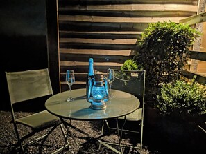 Ein Tisch mit einer blauen Laterne, zwei Gläsern und einem Stuhl in einer Gartenumgebung mit einer Holzverkleidung und viel Grün.