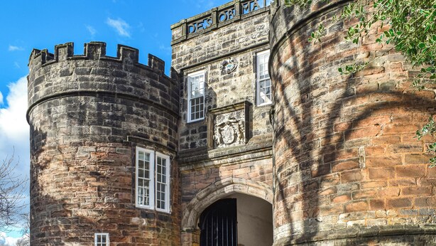 Een historisch gebouw van natuursteen met een ronde toren, een gebogen entree en een familiewapen.