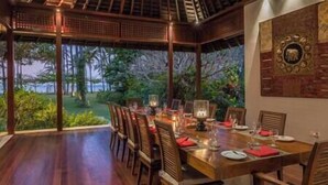 A dining area with a wooden table set for six, surrounded by chairs, with a view of greenery and a building in the background.