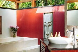 A bathroom with a white sink, a large mirror, and a decorative mask on the wall. - The Anandita by Elite Havens (Lombok Utara)