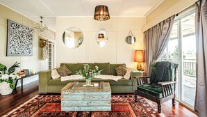 A living room with a green sofa, a wooden coffee table, and a patterned rug. There are two chairs, a chandelier, and a large window with curtains. - Charming 1940's two bedroom cottage, established gardens, large verandah and BBQ (Dorrigo)