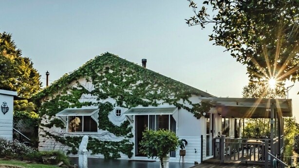 A white house with ivy-covered walls, a wooden deck, and a stone statue in the front yard. - Charming 1940's two bedroom cottage, established gardens, large verandah and BBQ (Dorrigo)
