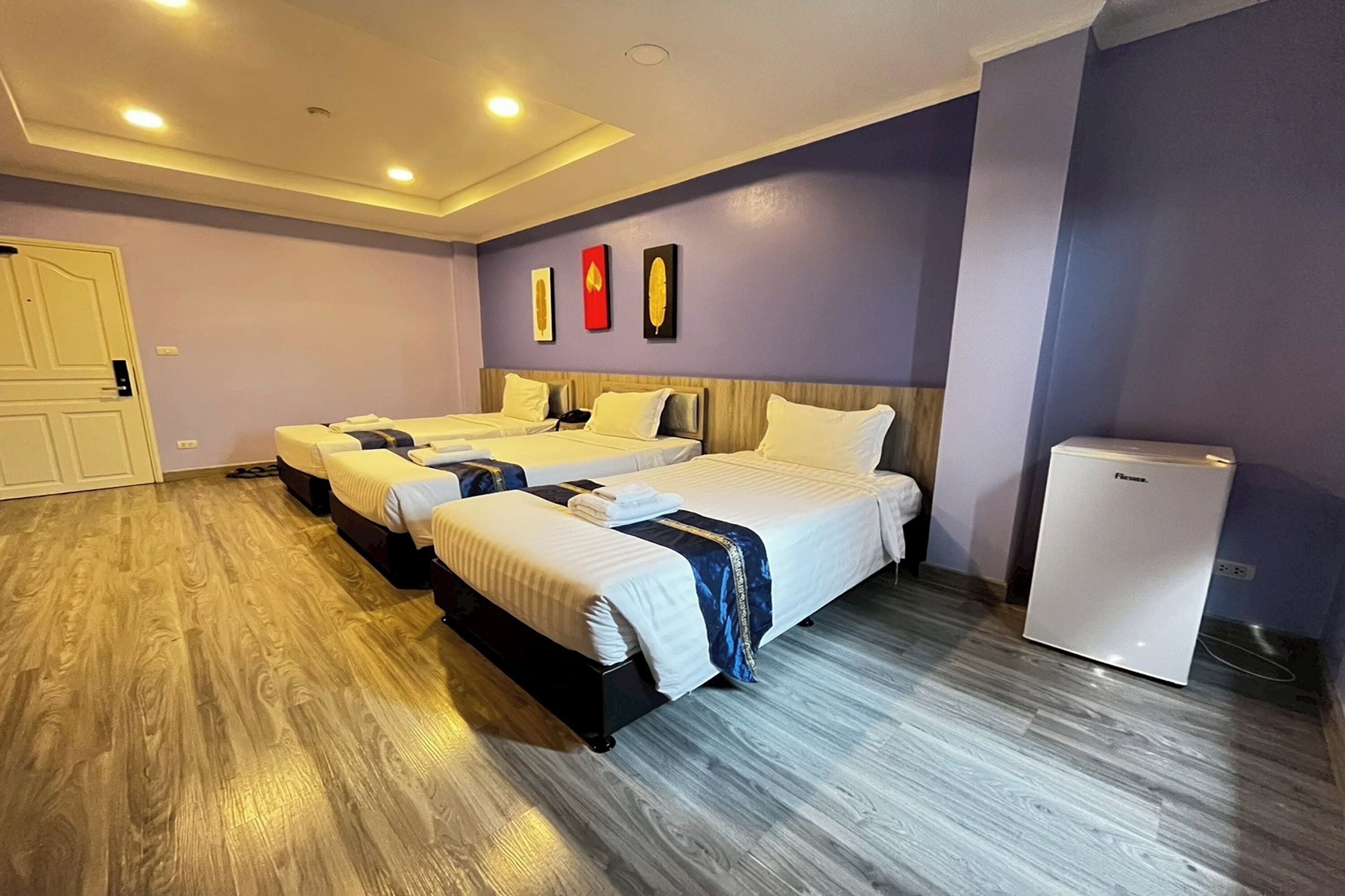 Une chambre d’hôtel avec trois lits, un réfrigérateur, un plancher en bois et une porte.
