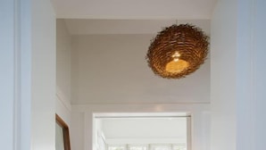 Um corredor com piso de madeira, um sofá branco com almofadas, uma luminária pendente de vime e uma porta branca que dá para outro quarto.