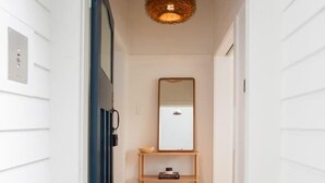 Um corredor estreito com piso de madeira, uma porta azul, um espelho em uma prateleira e um lustre.