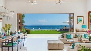 Amplia sala de estar con un ventanal que da a la playa y al océano. Hay un comedor con mesa y sillas, y un sofá con cojines.