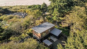 A modern cabin with a sloped roof, surrounded by dense forest and overlooking a beach. - CoHo Vista: Curated Comfort, High-End Style & Breathtaking Coastal Views, Seconds Away From Beach (Rockaway Beach)