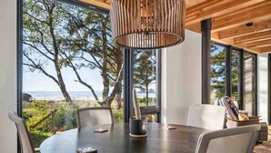 A round dining table with chairs, a hanging light fixture, and a view of the ocean through large windows. - CoHo Vista: Curated Comfort, High-End Style & Breathtaking Coastal Views, Seconds Away From Beach (Rockaway Beach)