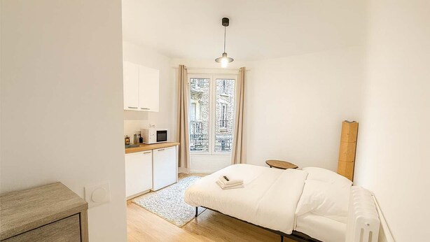 A compact hotel room with a bed, a small kitchenette, a window with a view, and a wooden headboard. - Studio Paris 15ème: Proche Métro Parfait pour 2 (Paris)