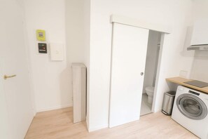 A compact laundry room with a washing machine, a trash bin, and a small wooden shelf.