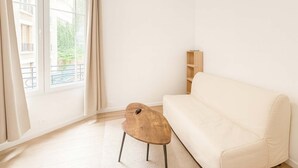 A room with a white sofa, a small wooden table, and a window with curtains. - Studio Paris 15ème: Proche Métro Parfait pour 2 (Paris)