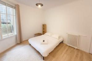 A bedroom with a bed, a small wooden nightstand, a radiator, and a window with curtains.