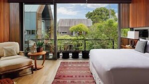 Une chambre avec un grand lit, une chaise, une petite table avec une plante, et une vue sur une maison et la verdure, visible à travers une porte vitrée.