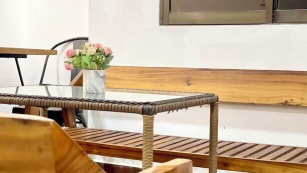 A wooden bench with a woven backrest, a white vase with pink flowers, and a window with a metal frame. - SKY Apartments - 50m to National Museum (Phnom Penh)