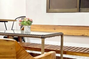 Un banc en bois avec un dossier en osier, un vase blanc orné de fleurs roses et une fenêtre dotée d’un cadre en métal.