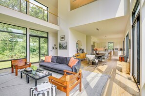 A spacious living area with a sofa, coffee table, and chairs, large windows offering a view of greenery, and a kitchen in the background. - Hudson Valley Summer Escape w/ Pool & Fire Pit (Pine Plains)