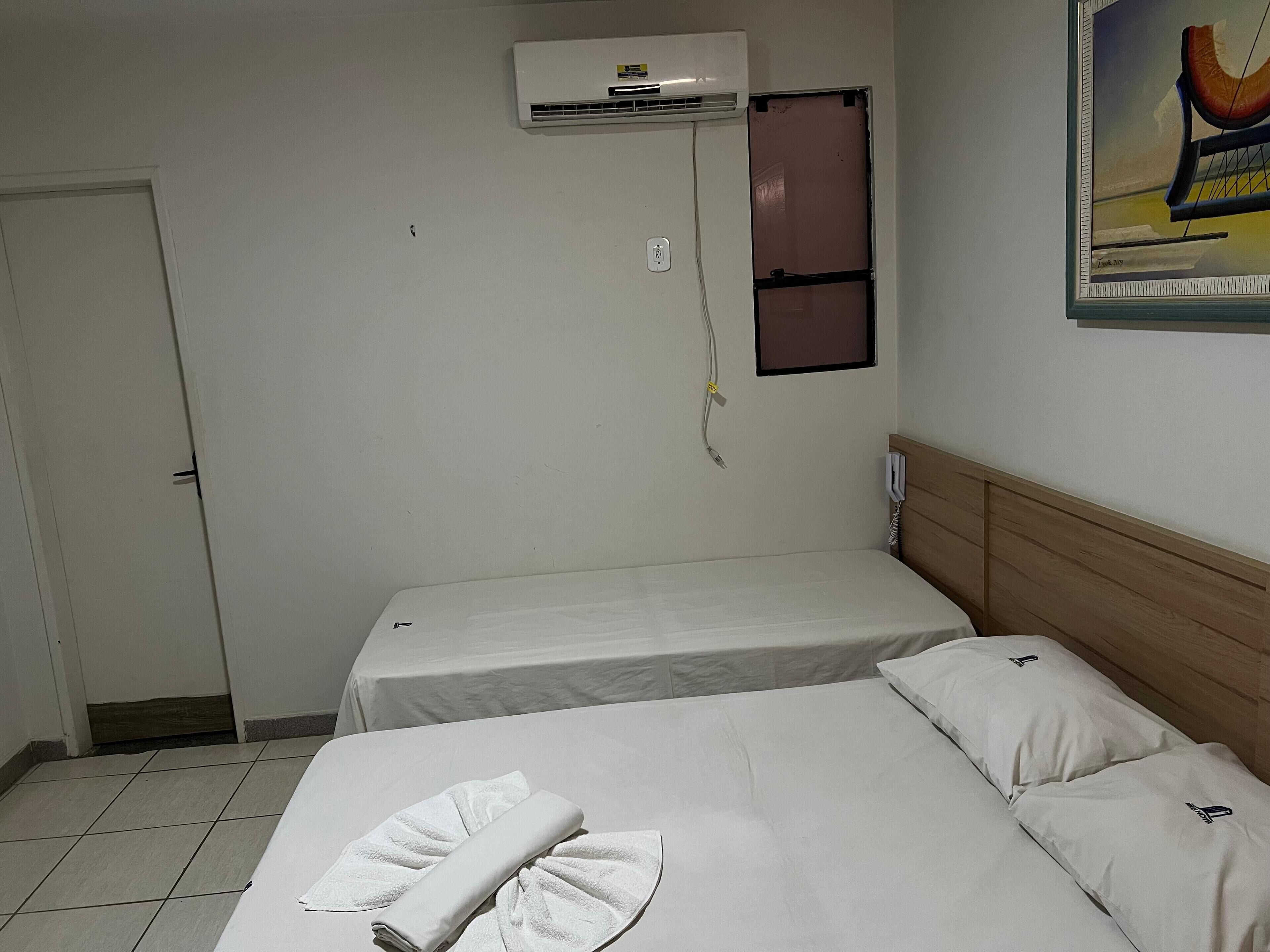 Een hotelkamer met een bed, nachtkastje, airconditioning en een schilderij aan de muur.