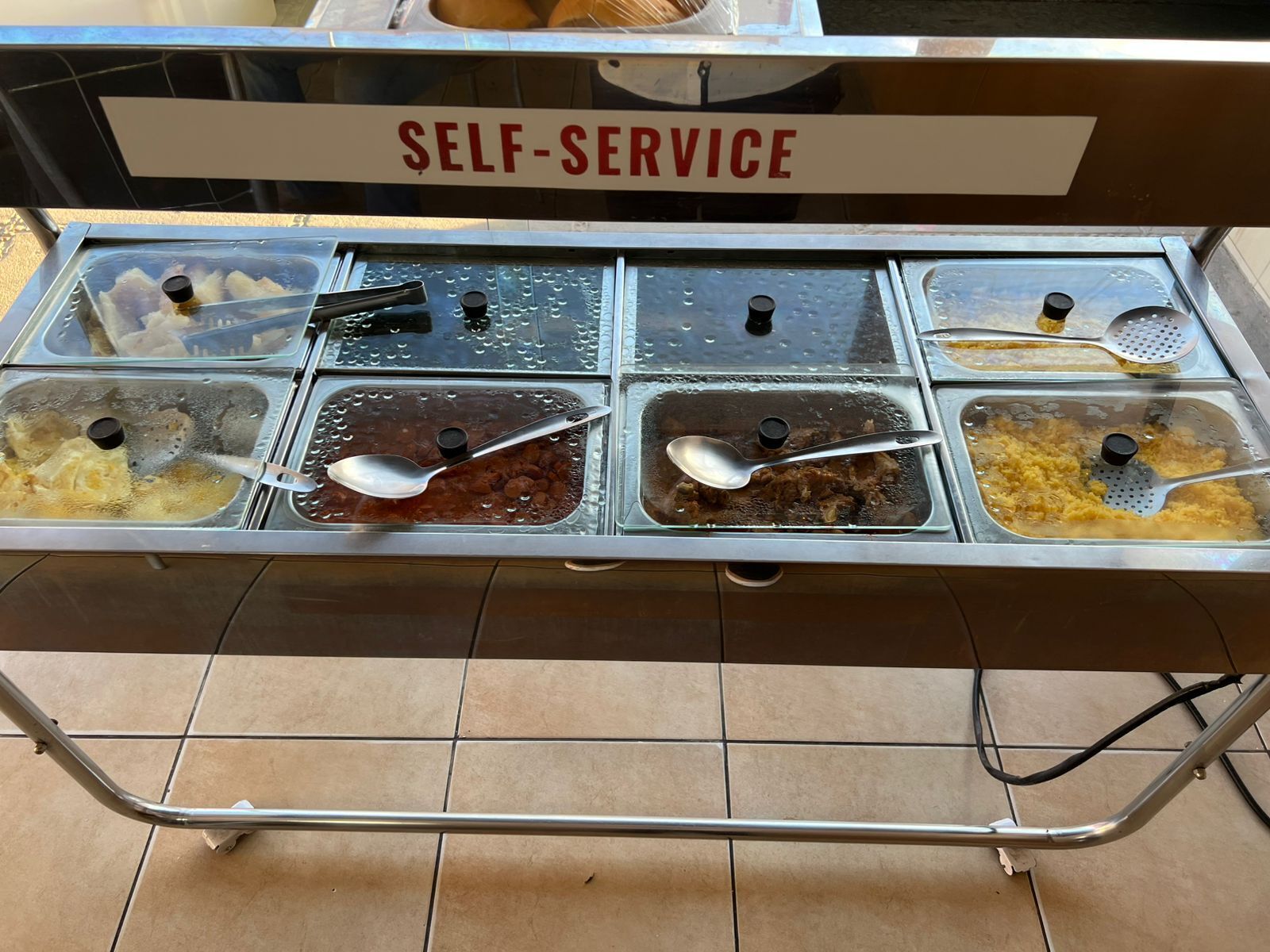 A self-service food station with multiple trays of food and spoons.