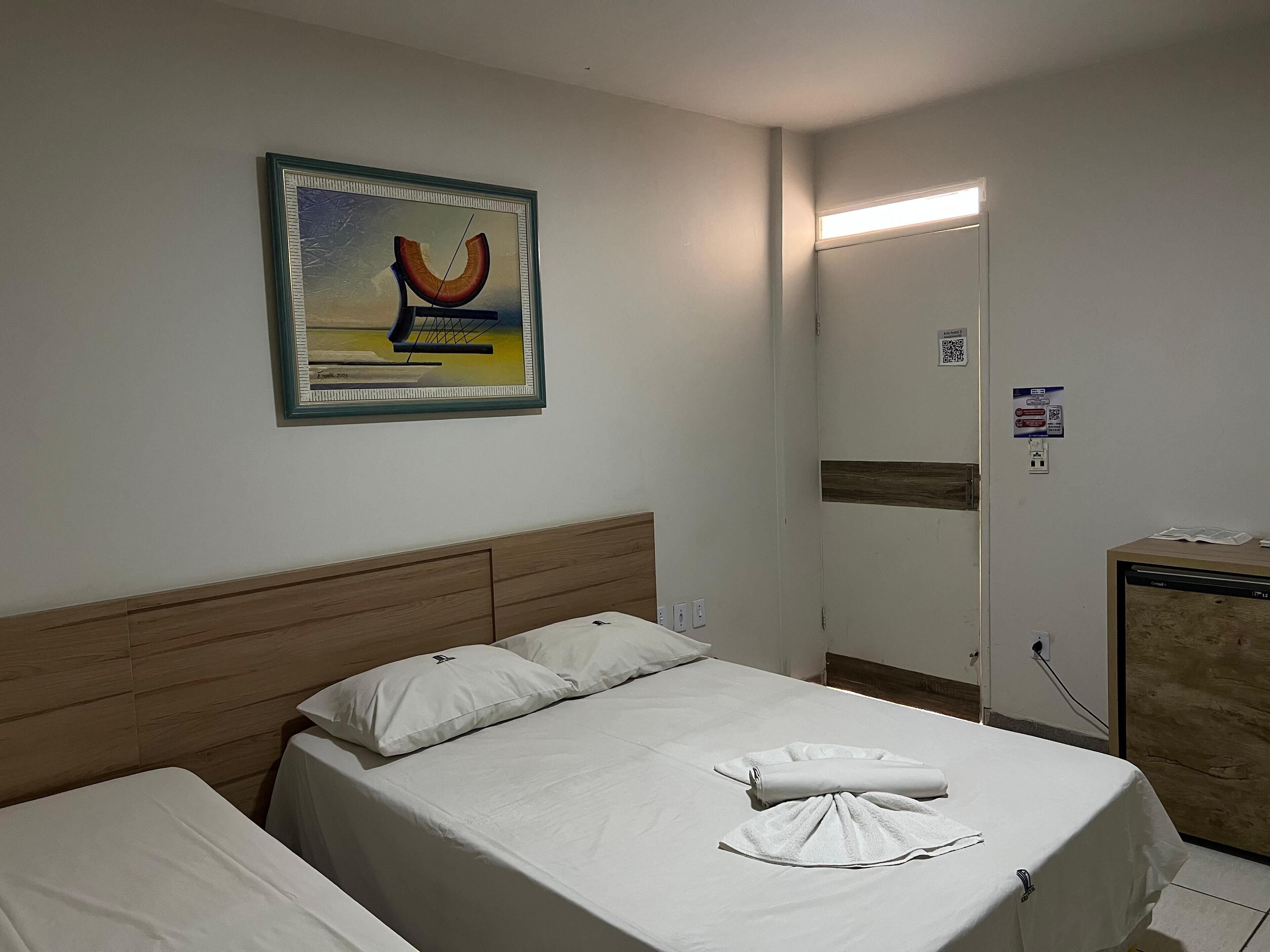 A hotel room with a bed, a headboard, a painting, a mirror, and a desk with a computer.