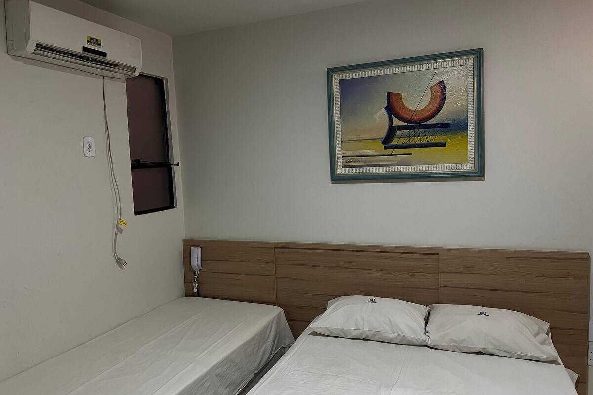 A hotel room with a single bed, a wooden headboard, an air conditioning unit, a power outlet, and a framed picture on the wall.