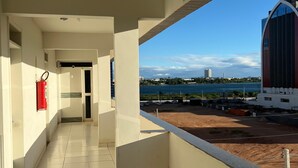 A corridor with a fire extinguisher on the wall, leading to a door. A sign indicates an exit. The corridor has a tiled floor and a view of a building and a body of water in the distance. - Kris Hotel (Petrolina)