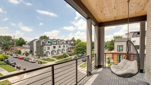 A balcony with a hanging chair, a view of a multi-story building, and a street with parked cars.