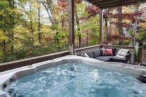 A hot tub with a wooden deck and outdoor furniture, surrounded by trees with autumn foliage.