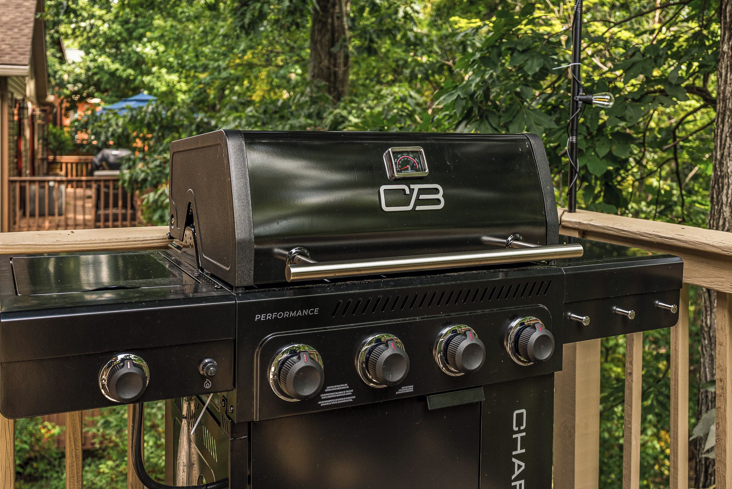 Ein CB Performance Grill mit vier Brennern und einem Seitenbrenner auf einer Holzterrasse mit Blick auf einen Garten.