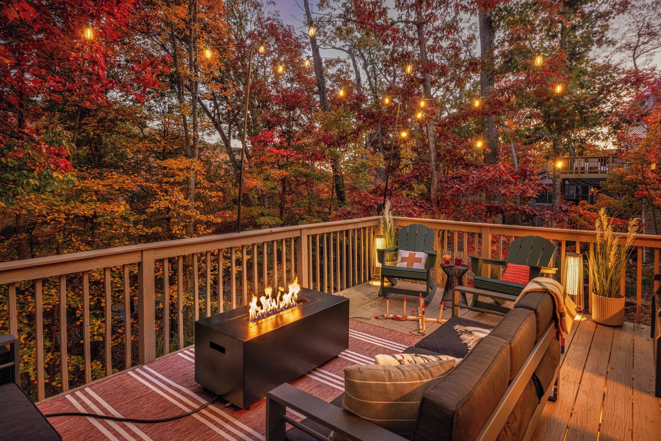 A deck with a fire feature, seating, and string lights against a backdrop of autumn foliage.