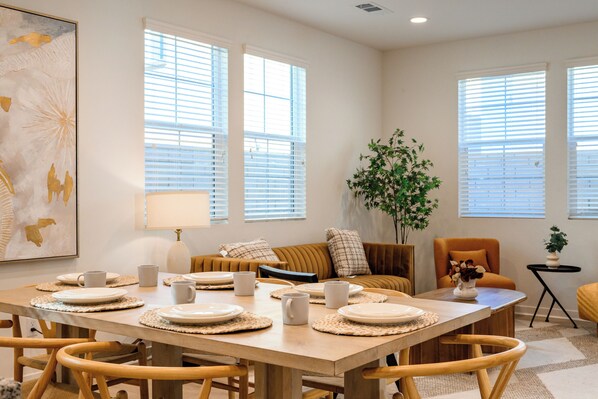 Un comedor con una mesa de madera dispuesta para seis personas, rodeada de sillas de madera. Hay dos ventanas con persianas, un sofá con cojines y una pequeña mesita con una planta.