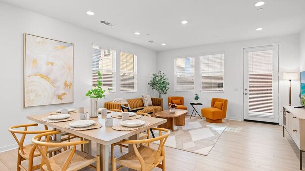 A spacious living area with a dining table set for four, surrounded by wicker chairs. There is a sofa and two armchairs, a coffee table, and a potted plant.