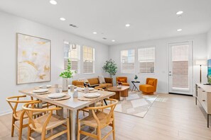 Amplia sala de estar con una mesa de comedor preparada para cuatro personas, rodeada de sillas de mimbre. Hay un sofá y dos sillones, una mesa de centro y una planta en maceta.
