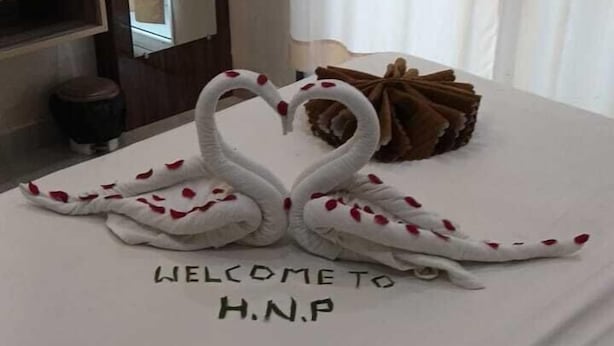 A neatly made bed with a swan towel design, a welcome sign, and a rose decoration. - Hotel Naman Palace (Varanasi)