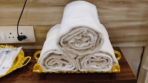A neatly folded white towel on a golden tray, with a power outlet and a cable in the background. - Hotel Naman Palace (Varanasi)