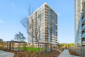 Un immeuble moderne de grande hauteur, doté de nombreux balcons, entouré d’un jardin aménagé avec des allées et des espaces de repos.