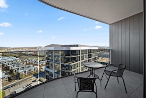 A balcony with a view of a modern building complex, two chairs, and a small round table.